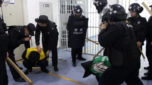 Заключенный сидит на корточках, опустив голову, в окружении офицеров с дубинками